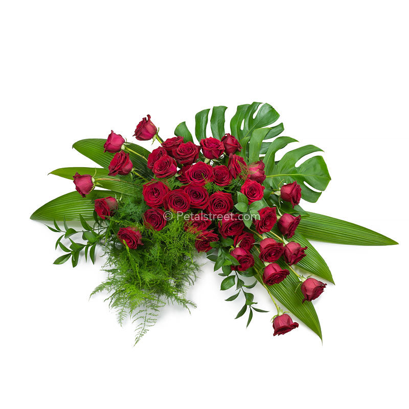 Stylishly designed red Rose Casket Spray with Feather Fern and Ruscus accents, and Accordion Leaves, and Monstera Leaves.