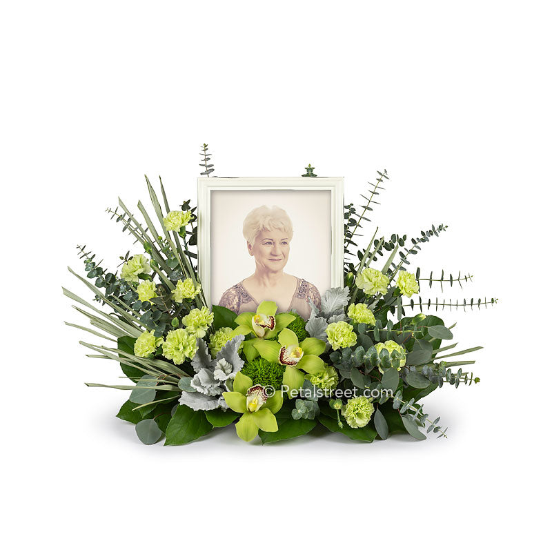 This cremation urn flower arrangement (shown with tribute photo frame) includes Cymbidium Orchids, mini Carnations, Green Trick, Eucalyptus, and accent foliage.