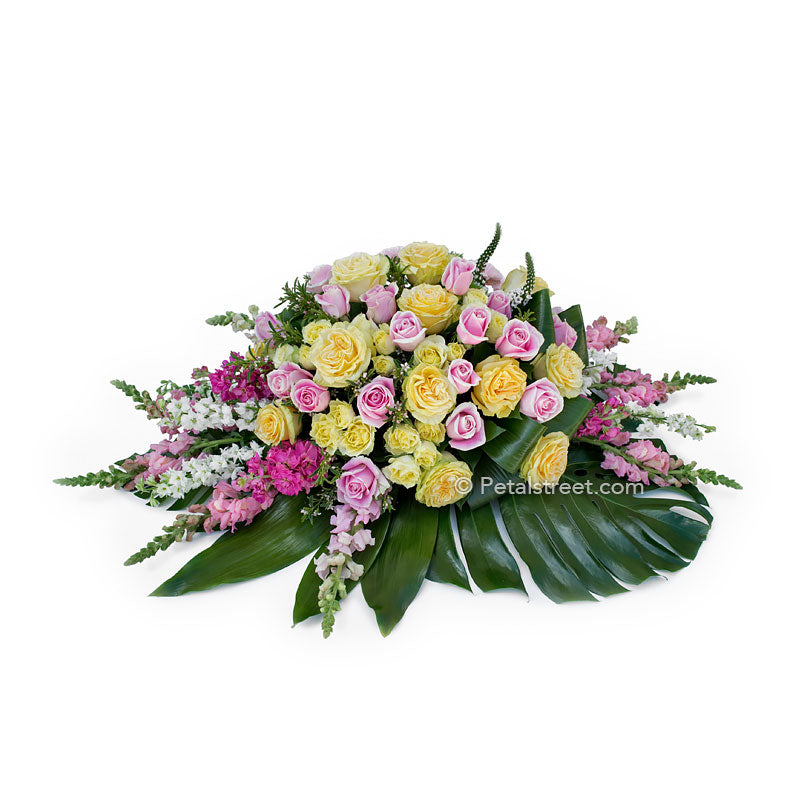 Beautiful artisan styled casket spray with pink and yellow Roses, Snapdragons, and large Monstera leaves.