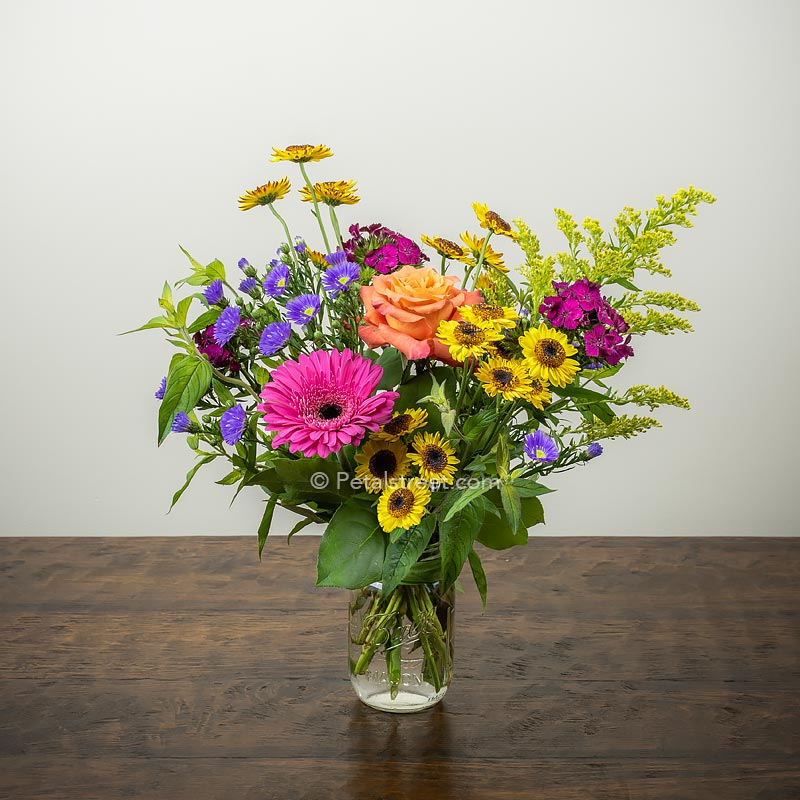Vibrant mix of flowers such as Gerbera Daisies, Roses, Viking Poms, Solidago, Aster, and accent greenery in a mason jar.