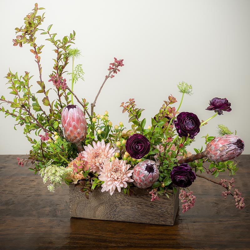 Beautiful bohemian flower arrangement with soft colors in a rustic wood garden box featuring Protea, Ranunculus, Lace Flower, Mums, and accents branches at Petal Street Flower Company florist