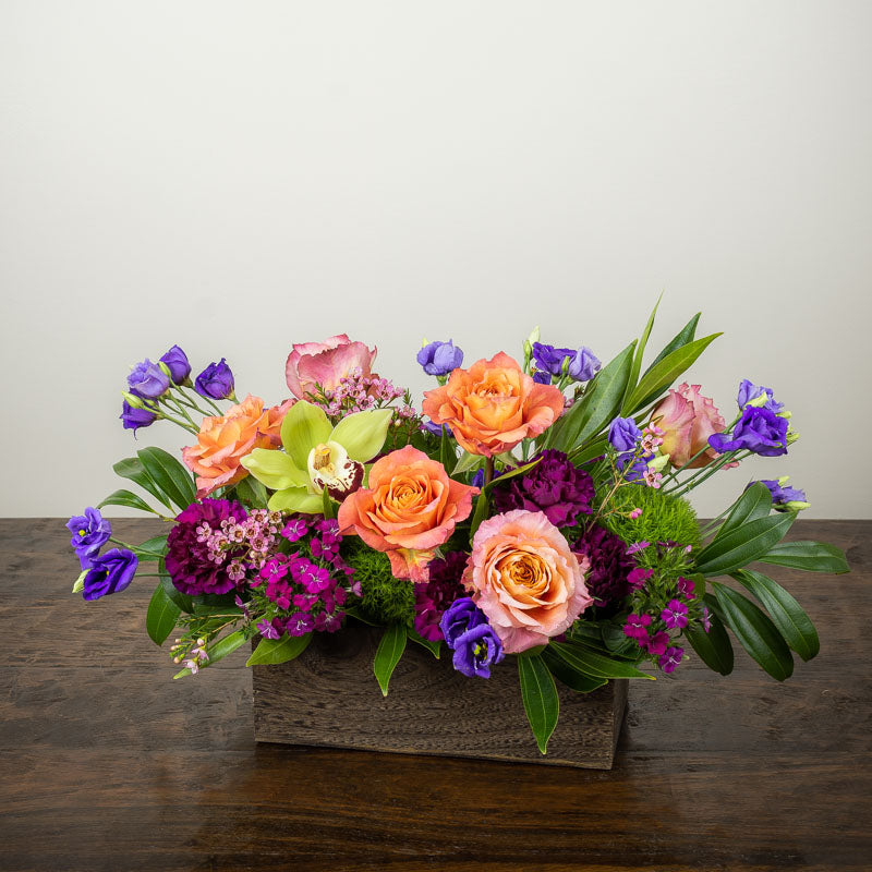 Colorful mix of flowers arranged in a wood box with orange roses, green Cymbidium Orchids, purple Lisianthus, magenta Sweet William, with accent blooms and greenery
