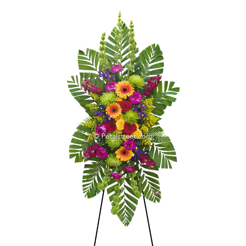 Colorful standing spray with yellow and orange Gerbera Daisies and Roses, floral accents, and lush greenery.