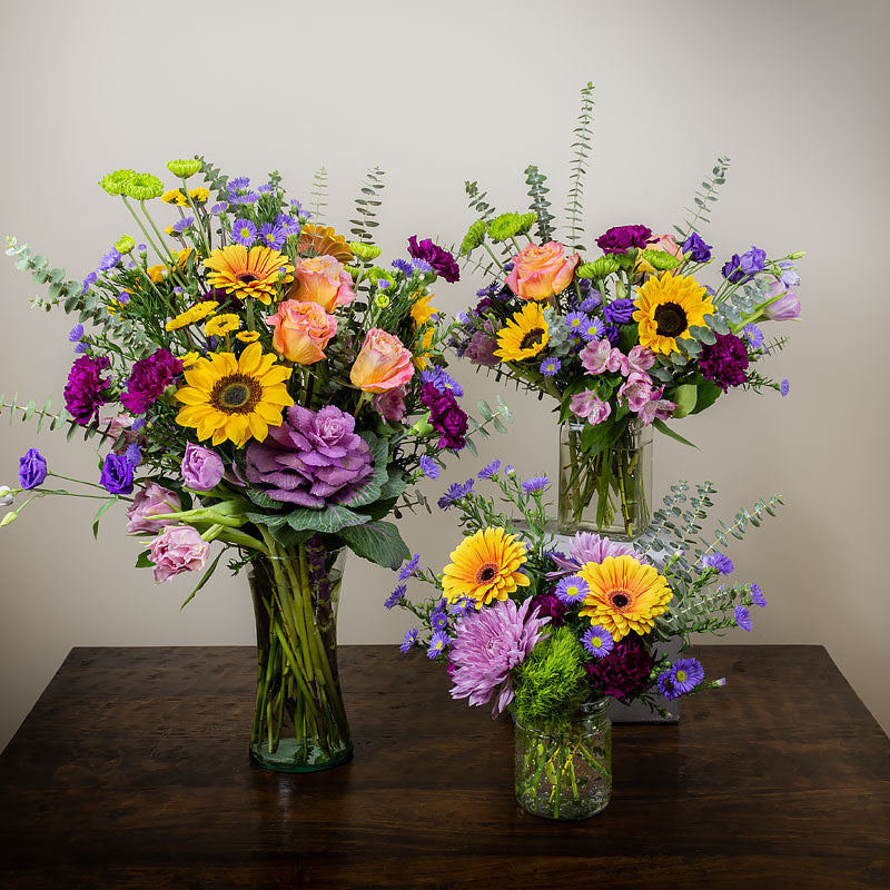 A group of three vibrant colored flower arrangements, small, medium, and large, featuring a variety of blooms such as Roses, Sunflowers, Gerbera Daisies, Mums, Eucalyptus, and accent greenery at Petal Street Flower Company florist