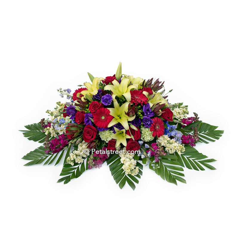 Bright and bold colored casket spray with lilies, Roses, Daisies, Snapdragons, and large leaf accents.