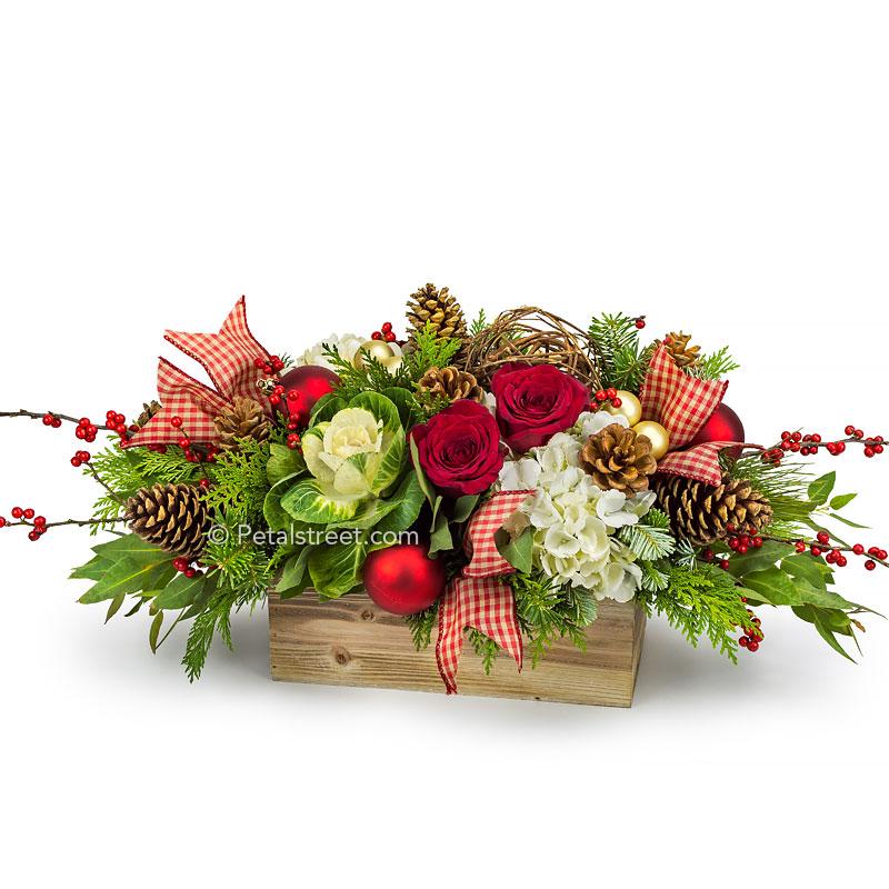Country Christmas styled flower arrangement with red Roses and Berries, green Kale, white Hydrangea, Pine Cones, red and gold holiday ornaments, plaid ribbon tails, and mixed seasonal greens arranged in a rustic wooden box.