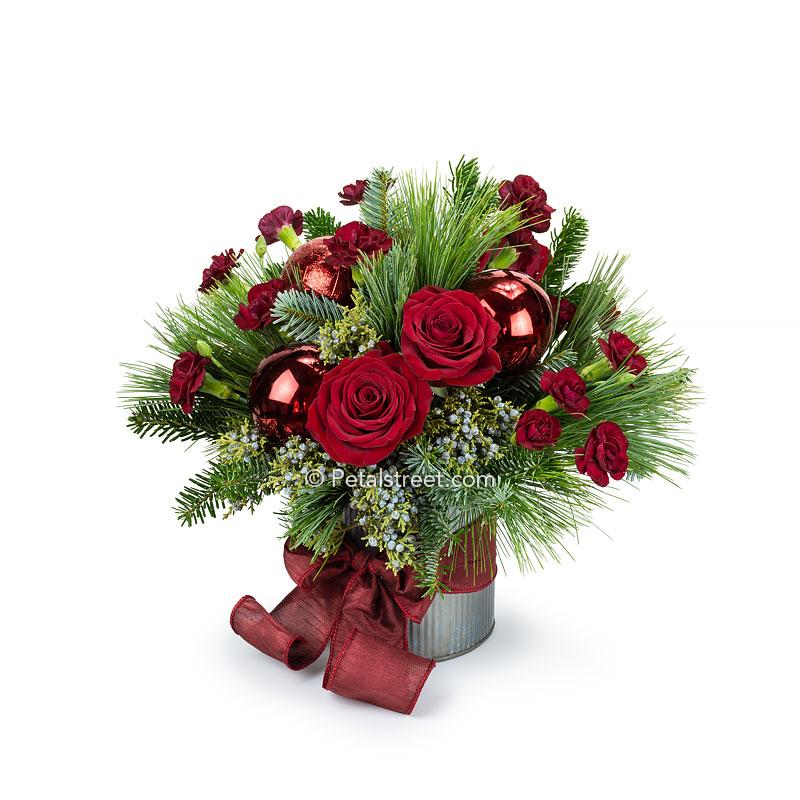 Christmas flower arrangement with red Roses, red mini Carnations, blue Juniper Foliage, mixed seasonal greenery, and large red holiday ornaments in a rustic tin cylinder container with a hand-tied maroon bow.
