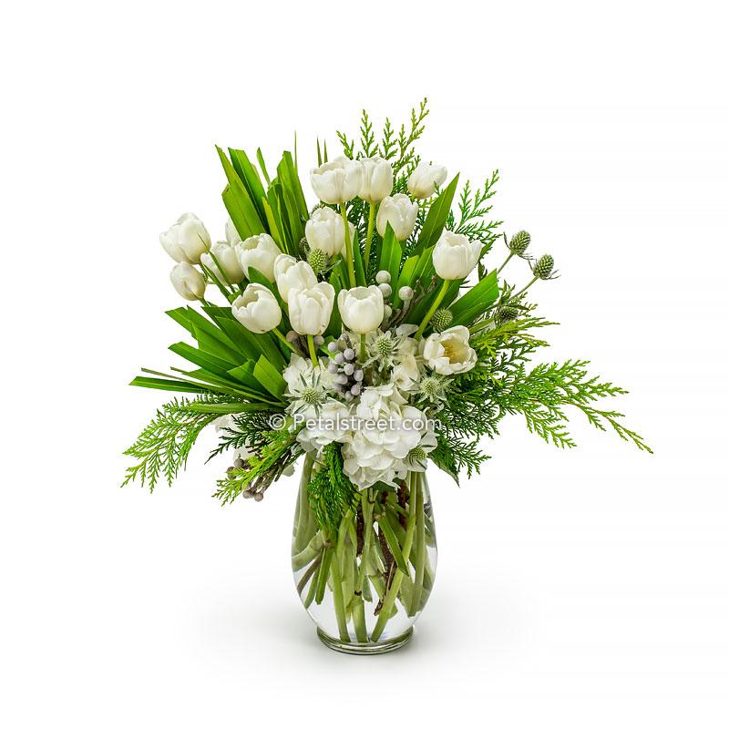Elegant white and green Christmas flowers in a vase with white Tulips, white Hydrangea, Thistle, Brunia, hand-cut Fan Palm, and Cedar greenery.