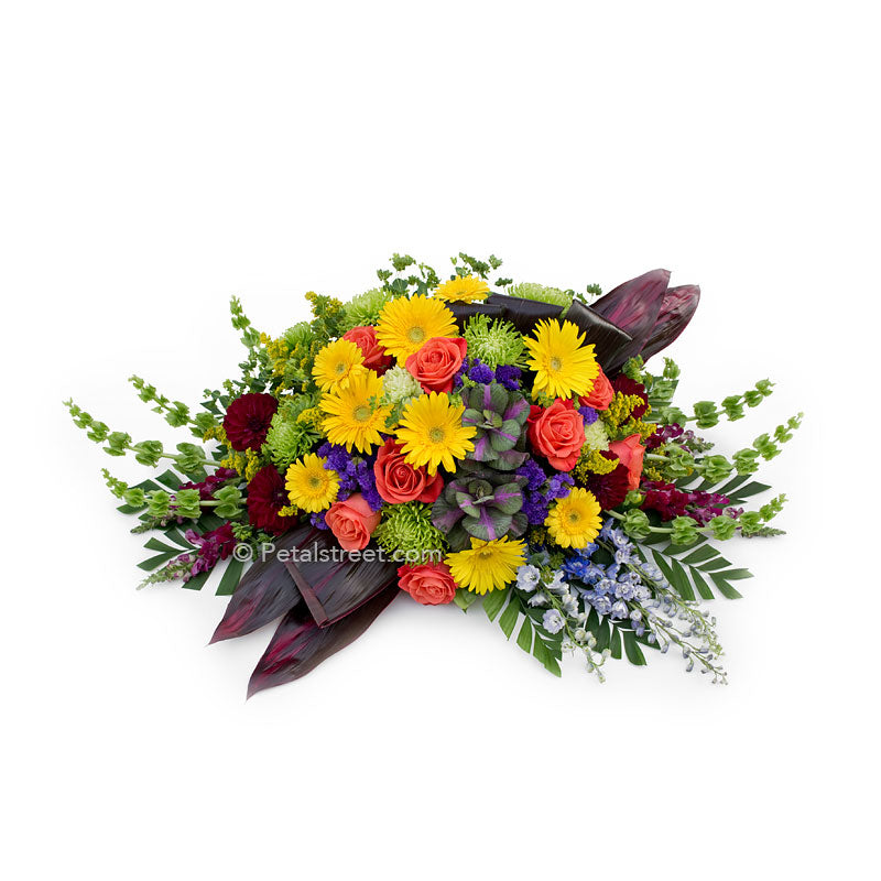 Bright colored casket spray with yellow Daisies, orange Roses, green Spider Mums, and deep earth toned Ti leaves.