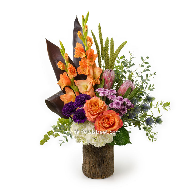 Orange Roses and Gladiolas, plum Carnations and Mums, Protea, Thistle, Hydrangea, and Millet arranged in a Bark cover vase with Ti Leaf accents.