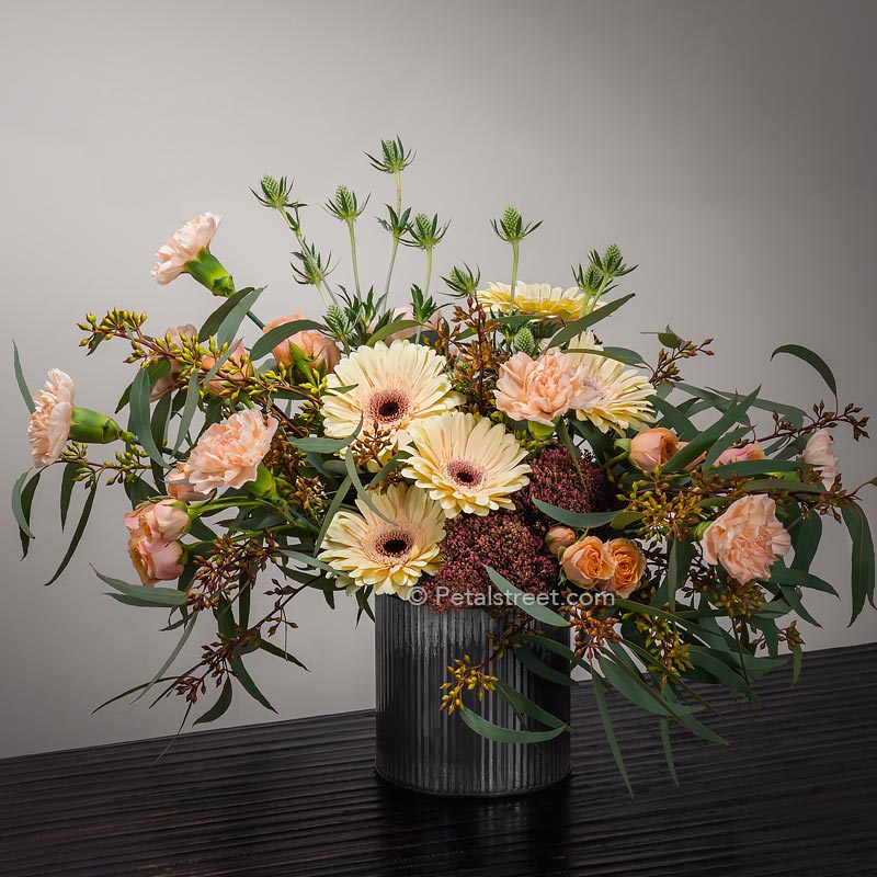 Soft peach and orange flowers such as Gerbera daisies, Carnations, and mini Roses, with Sedum, Eucalyptus and Thistle accents arranged in a zinc tin vase.