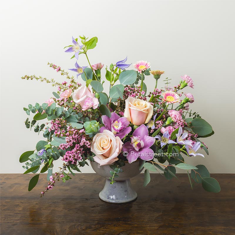 Gorgeous low wide mixed flower arrangement with soft peach Roses, Orchids, Clematis, and Eucalyptus.