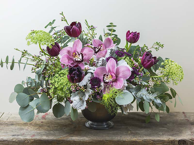 Sophisticated garden mix Spring Flower arrangement with pink Orchids and purple Tulips arranged in a low compote style container photographed on a wood table top