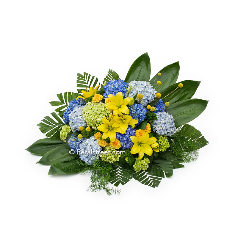 Modern floral designed casket spray with yellow Lilies, large blue and green Hydrangea, and lush green Ti Leaves.