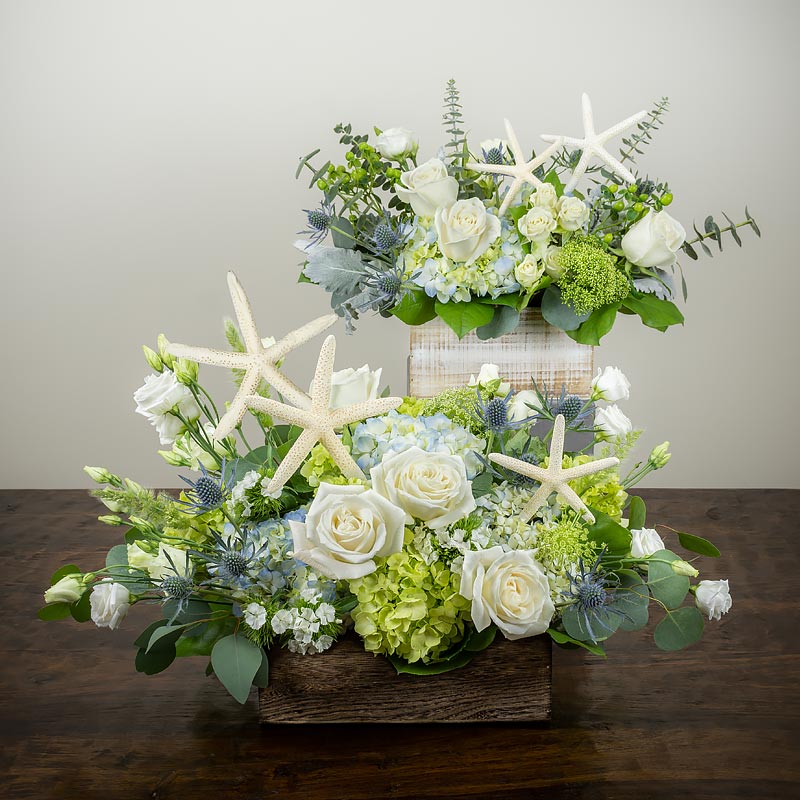 A pair of nautical or beach themed flower arrangements in wood garden boxes featuring white Roses and Lisianthus, blue and green Hydrangea, Eucalyptus, and starfish at petal Street Flower Company florist