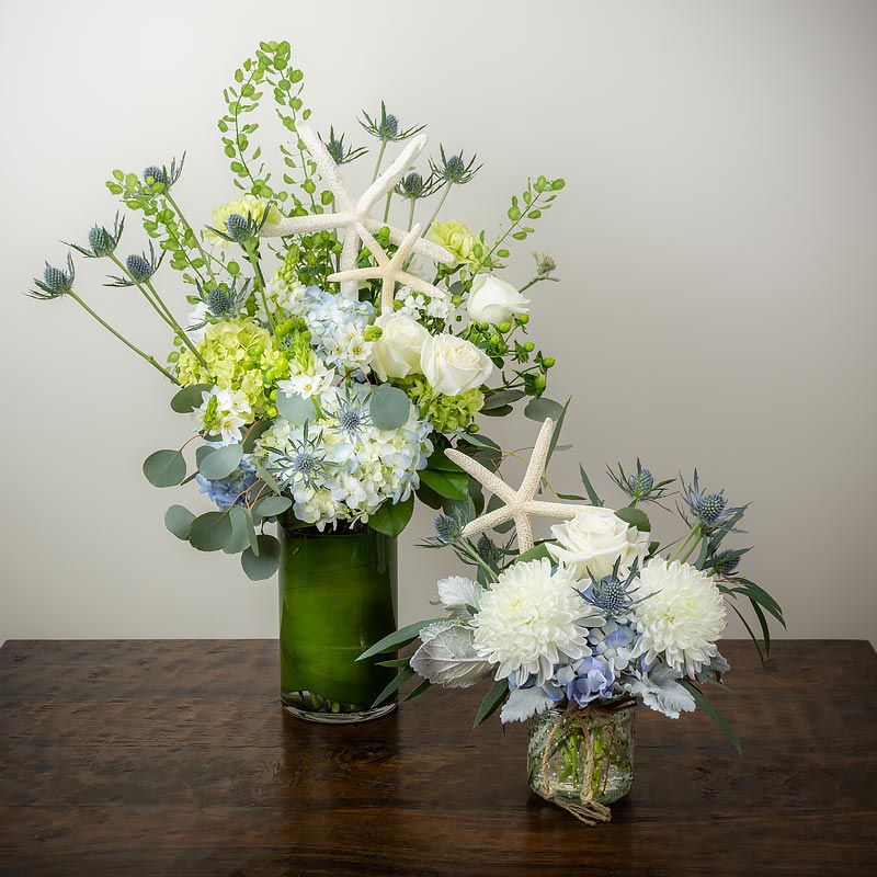 A pair of nautical or beach themed flower arrangements in vases featuring white Roses and Mums, Thistle, blue and green Hydrangea, Eucalyptus, and starfish at petal Street Flower Company florist