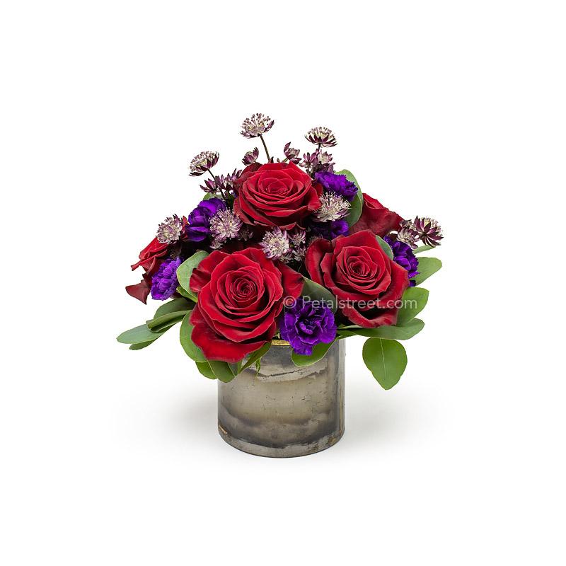 Contemporary styled vase of flowers with red Roses, purple mini Carnations, and accents arranged in a metal vase.