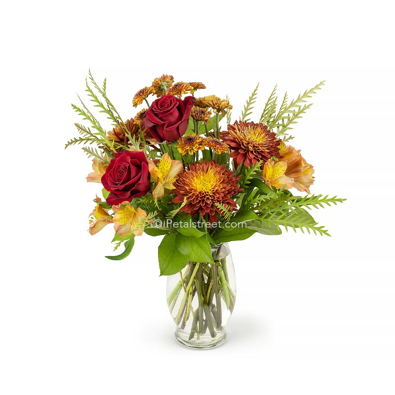 Mixed Autumn flower arrangement with brown orange mums, red roses, mini button mums, orange alstroemeria, and accent foliage in a vase.